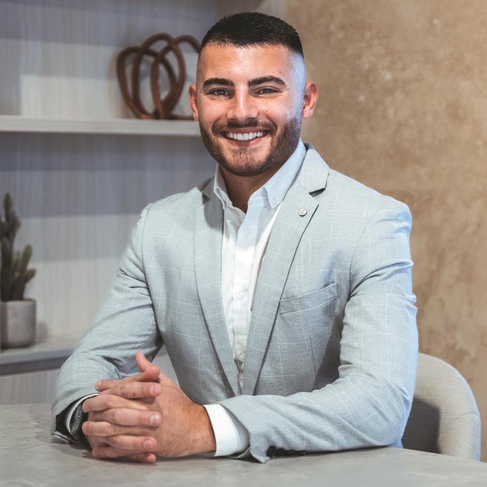 The image features a well-dressed individual seated at a modern desk. He is smiling, exuding professionalism and confidence, ideal characteristics for a plumber representing Penrith City Plumbing Services. The background features minimalistic decor with a shelf displaying decorative items and a potted plant, suggesting a tidy and welcoming office environment. The person's attire, a light grey suit with a white shirt, reinforces a sense of professionalism appropriate for client interactions. This setting could be where consultations or customer service occurs, highlighting the business's commitment to professionalism in its plumbing services.