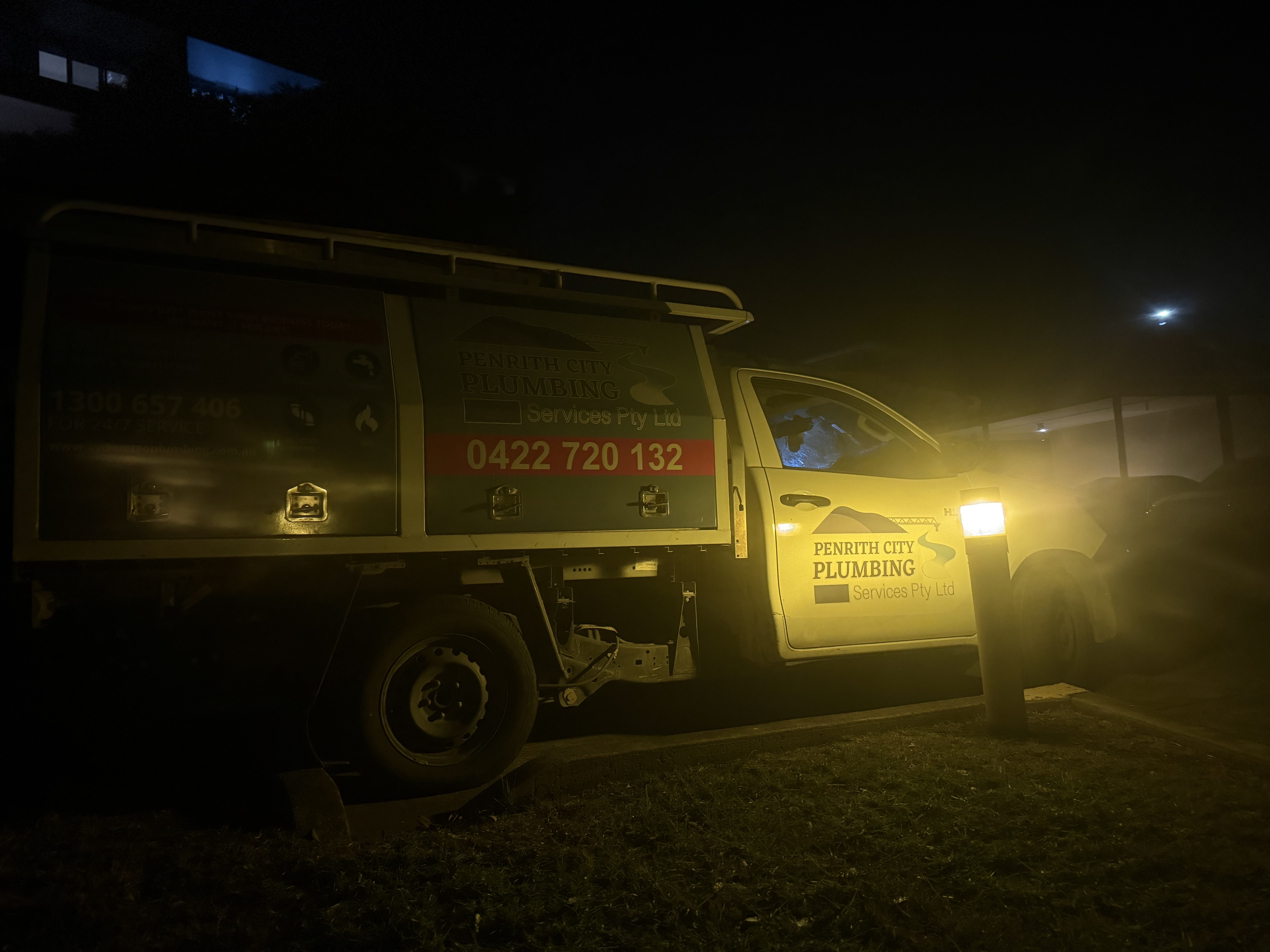 The image depicts a service vehicle belonging to Penrith City Plumbing Services, operating during the night. The truck is parked in a dimly lit area, illuminated by a streetlight and the vehicle’s own lights. A technician is visible inside, possibly assessing a job or preparing equipment for plumbing work. The vehicle displays the company’s branding prominently, which includes both the name and contact information. This suggests that the company is on-call and ready to attend to plumbing emergencies or scheduled maintenance in the Mulgoa area and surrounding locations. The nighttime setting indicates responsiveness and readiness to provide services, essential for a plumbing business that may operate outside regular hours. Overall, the image captures the professionalism and reliability of Penrith City Plumbing Services, essential in their line of work.