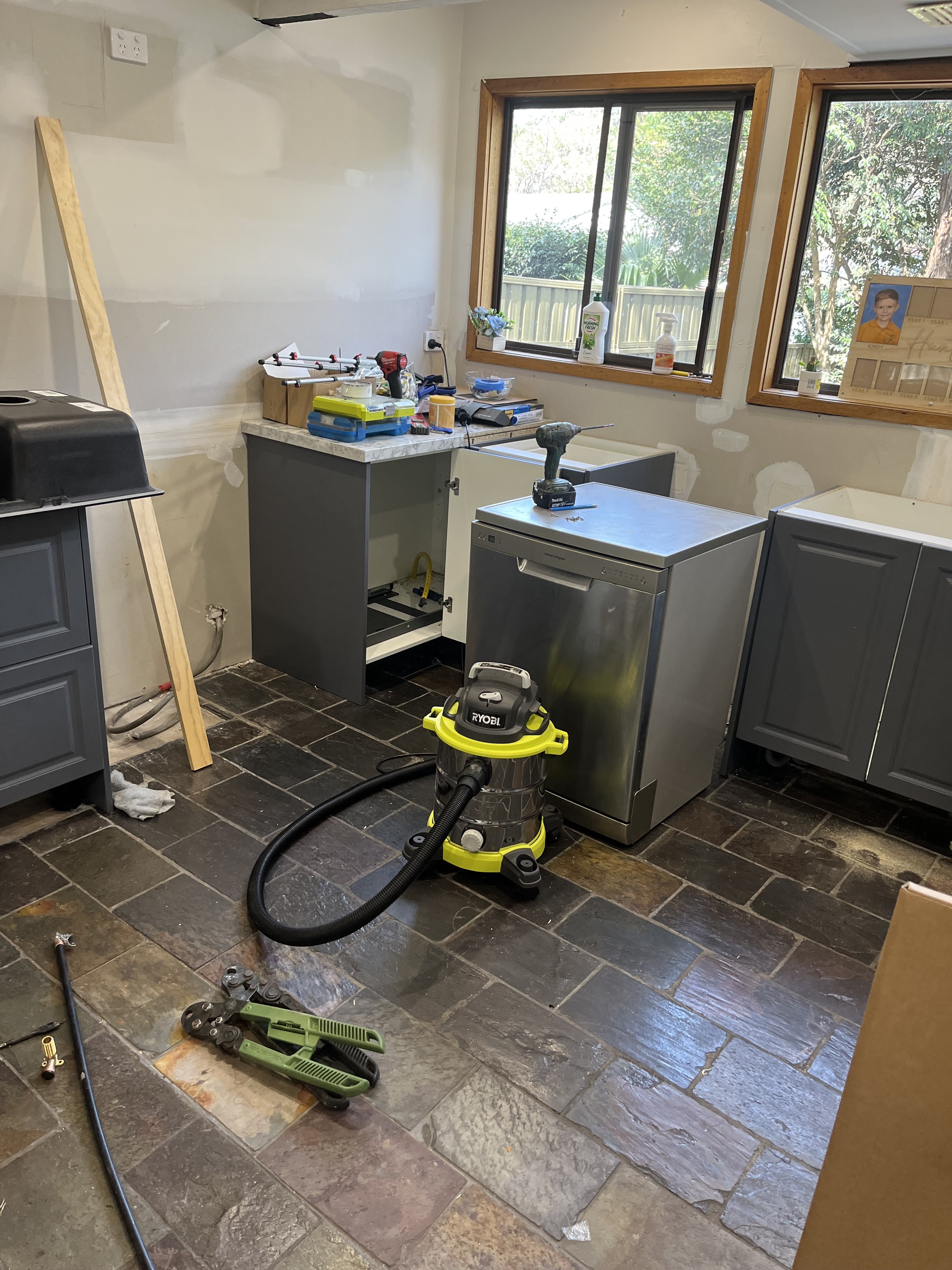 The image depicts an unfinished kitchen renovation scenario typical for a plumbing service environment. On the floor is a variety of equipment and tools, including a vacuum cleaner and a small tool kit, indicating ongoing plumbing work. The cabinets are recently installed, with one showing signs that it is ready for plumbing fixtures to be connected. The workspace appears cluttered, typical of renovation sites, with items scattered around including plumbing supplies and tools suggesting that the space is being prepared for future installations. The large window allows natural light into the room, highlighting the work that still needs to be completed.