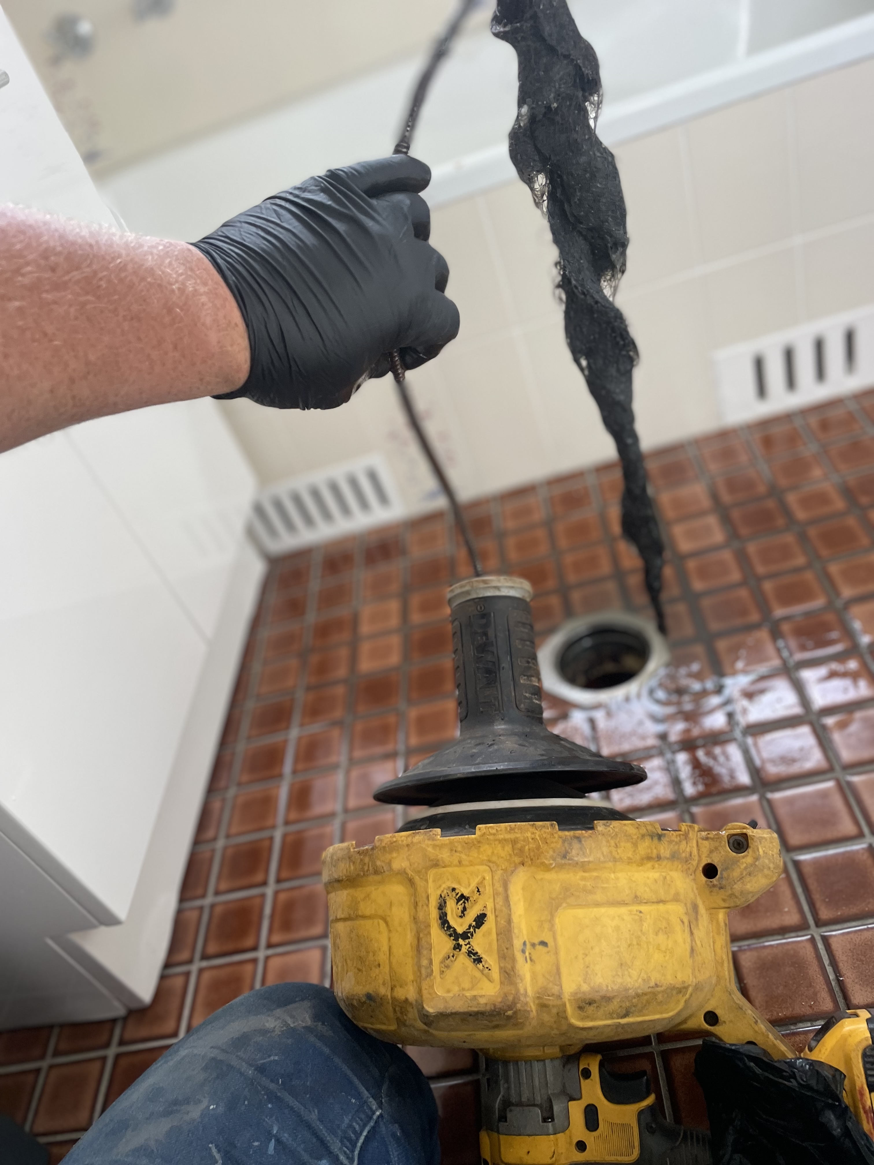The image shows a plumber's hand wearing a black glove, actively using a plumbing snake (auger) to remove a blockage from a drain. The yellow device appears to be an electric drain cleaner, which is often used to tackle stubborn clogs in pipes. The scene is set in a bathroom, evident from the tiles and drainage system visible in the background. A long, dark mass can be seen being extracted from the drain, likely comprised of hair and other debris. This indicates a routine maintenance or emergency plumbing service, showcasing Penrith City Plumbing Services' expertise in resolving plumbing issues in residential properties. Their prompt action in addressing such blockages is crucial for maintaining hygiene and function in the home, particularly in areas like Mulgoa and surrounding suburbs.