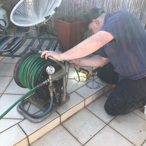 In the image, a plumber is working diligently with a high-pressure water cleaner, preparing to tackle a drain blockage or pipe problem. The plumber is crouched down beside a manhole cover, using a power drill to insert a plumbing snake or hose into the drain. The green hose is coiled and connected to the high-pressure cleaner, which suggests that a thorough cleaning or inspection is about to take place. The tiled surface indicates an outdoor setting, possibly a patio or backyard area. Nearby, there is a fan and potted plants, adding to the residential ambience. This scenario reflects typical plumbing services offered by Penrith City Plumbing Services in Mulgoa and surrounding areas, showing their readiness to address plumbing issues with professional equipment.