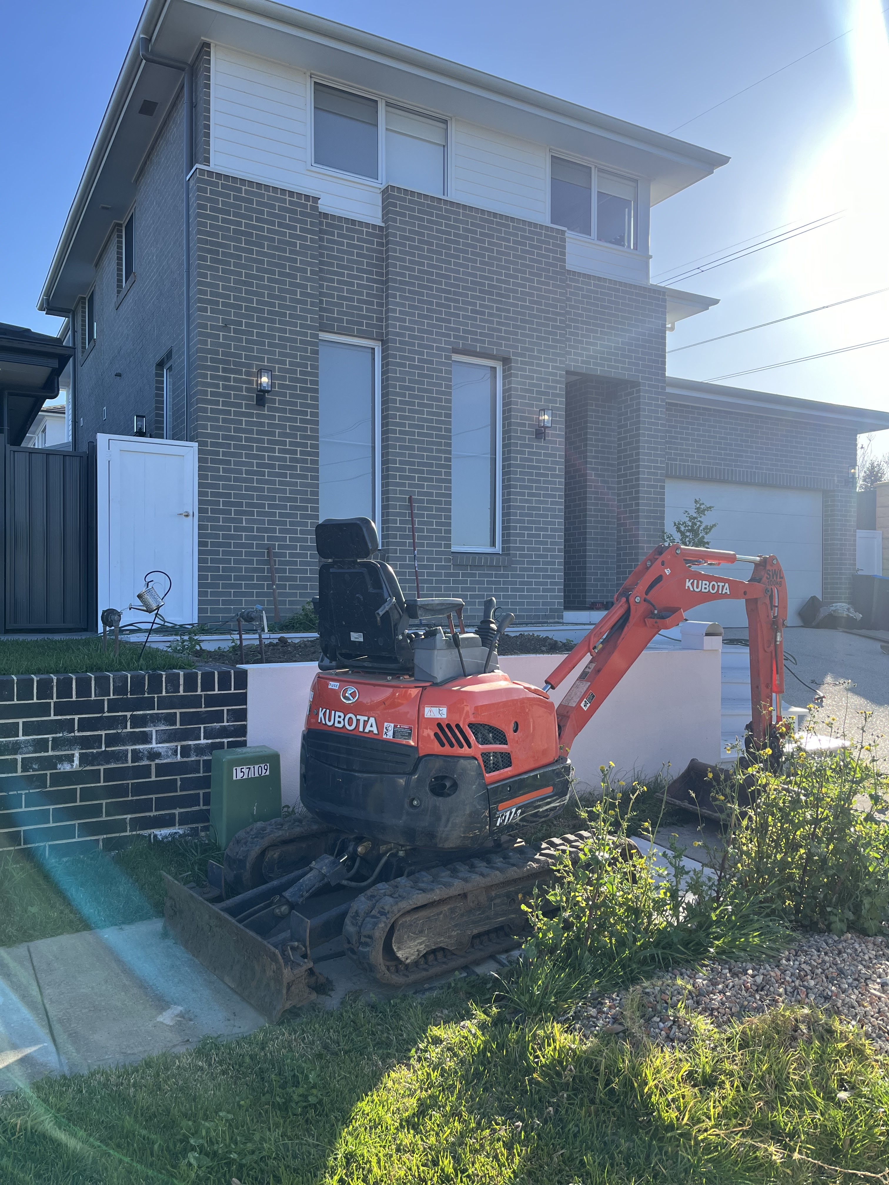 The image depicts a Kubota mini excavator parked on the driveway of a modern two-storey home. The excavator is likely being used for plumbing-related excavation work, such as digging trenches for pipe installation or repairs. The house features a contemporary design with a brick facade and large windows, showcasing a well-maintained exterior. With the excavator in place, it indicates that Penrith City Plumbing Services may be addressing plumbing issues at this residential property in Mulgoa, NSW, potentially relating to water supply or drainage systems. The presence of the excavator signifies that the plumbing work may involve substantial ground excavation, typical for plumbing services in both new construction and renovation projects.