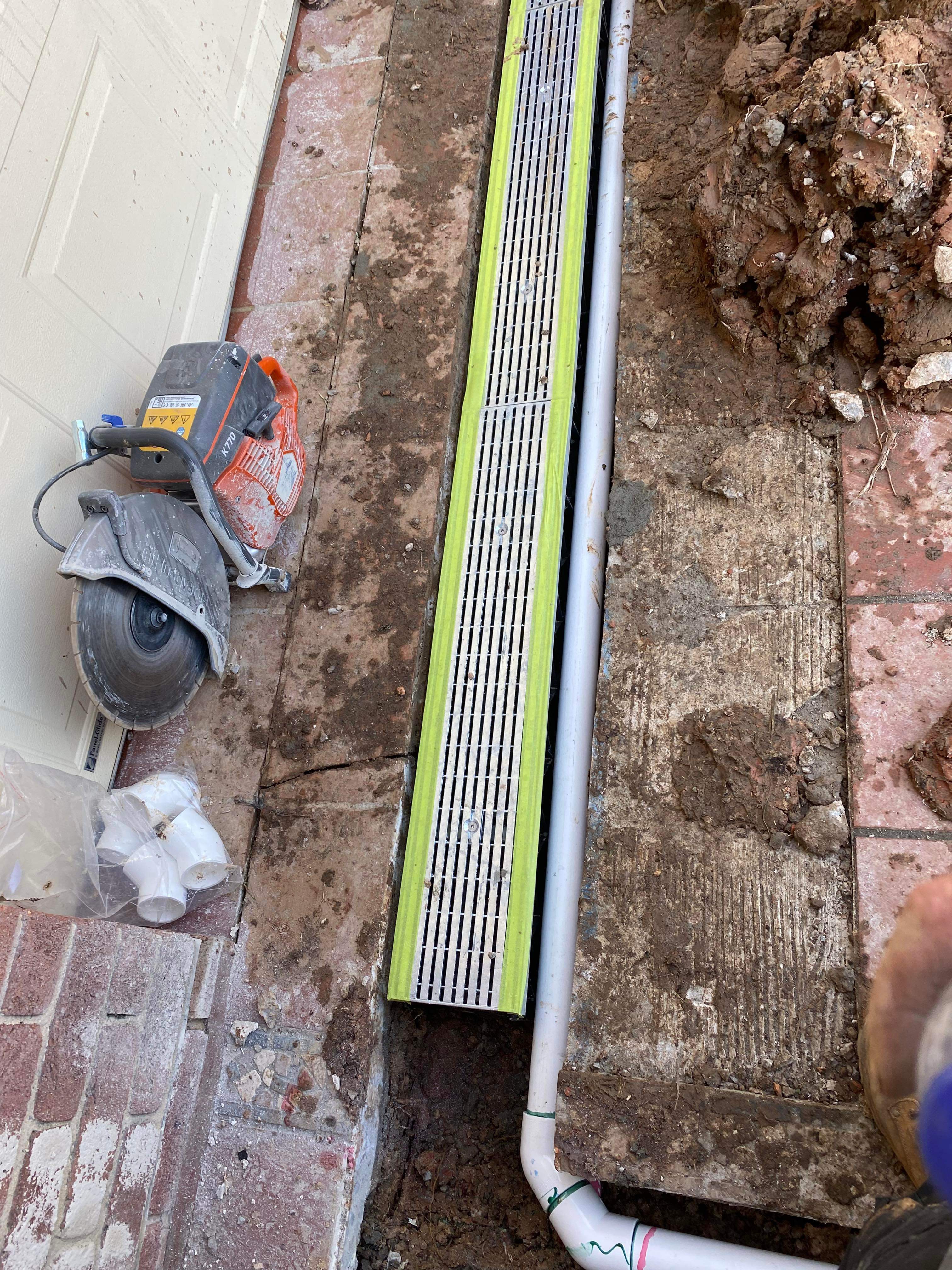 The image showcases a plumbing setup in a ground area, featuring a metal drainage grate that has been installed over a trench. The trench appears to be newly excavated and is adjacent to a paved surface, indicating an ongoing drainage project, likely aimed at managing water runoff effectively. A concrete saw is visible nearby, suggesting that cutting into pavement or other materials may be part of the project. Beneath the grate, white PVC pipes are arranged, implying that a drainage system is being connected, showcasing the functional aspect of plumbing work by Penrith City Plumbing Services. The presence of mud and dirt around the work area indicates it's an active construction site, where proper drainage solutions are being implemented to prevent water accumulation and potential flooding issues in the surrounding areas.