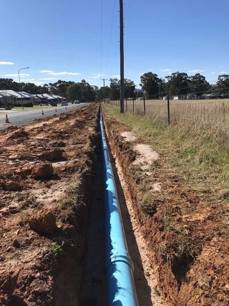 The image depicts a trench that has been dug in a rural area, likely in Mulgoa, NSW, with a long blue pipe laid along its length. The trench, which appears to have been excavated for plumbing or drainage purposes, is flanked by freshly disturbed soil. There are also power lines visible in the background, along with distant residential houses. The presence of construction cones indicates ongoing work, possibly related to upgrading or installing a pipeline system. This installation might be part of Penrith City Plumbing Services' efforts to improve water supply or drainage in the local area, ensuring efficient water management and infrastructure development to cater to the community's needs.