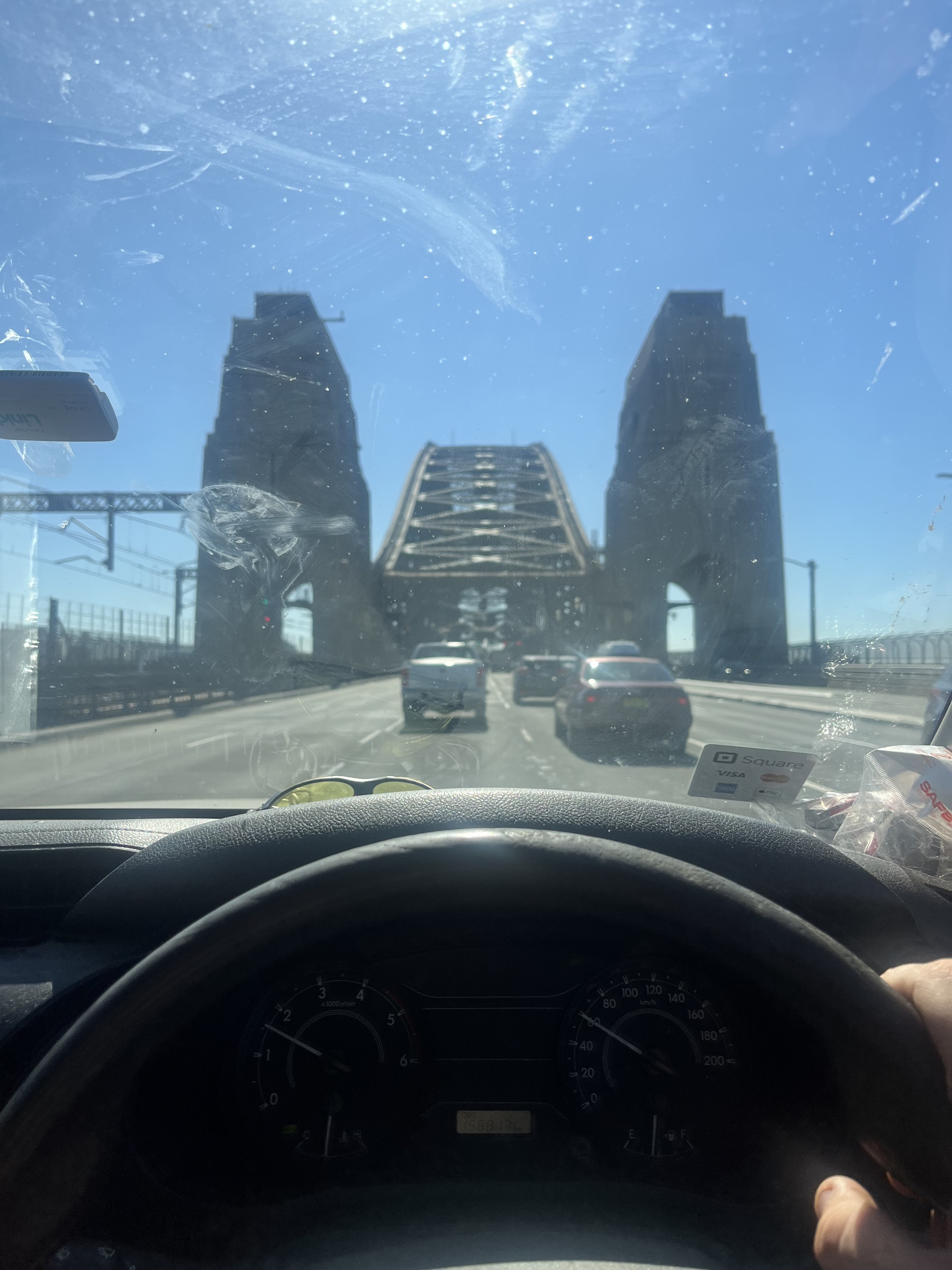The image captures a view from inside a vehicle, with the steering wheel prominently displayed in the foreground. In the background, the iconic Sydney Harbour Bridge is visible, showing its distinctive structure against a clear blue sky. The scene suggests a journey through Sydney, likely en route to a plumbing service call in one of the suburbs. This reflects the mobility and responsiveness of Penrith City Plumbing Services as they navigate the city, emphasizing their commitment to reaching clients across various locations including Mulgoa, Camden, and beyond. The presence of other vehicles indicates a busy day, hinting at the demand for reliable services in the area.