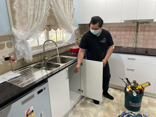 In the image, a plumber from Penrith City Plumbing Services is inspecting a kitchen sink area in a residential home. The plumber is wearing a black shirt and a face mask for safety, in line with health protocols. In the background, there is a modern kitchen layout with new cabinetry and dual stainless steel sinks, indicating ongoing renovation or installation. The plumber is opening a cupboard below the sink, likely checking for plumbing connections or potential leaks. Tools are visible in a bucket nearby, suggesting that he is prepared for various tasks related to plumbing, such as repairs or installations. The setting reflects a typical plumbing job, focusing on maintaining or enhancing the functionality of the home's water system, which is aligned with the services Penrith City Plumbing offers.