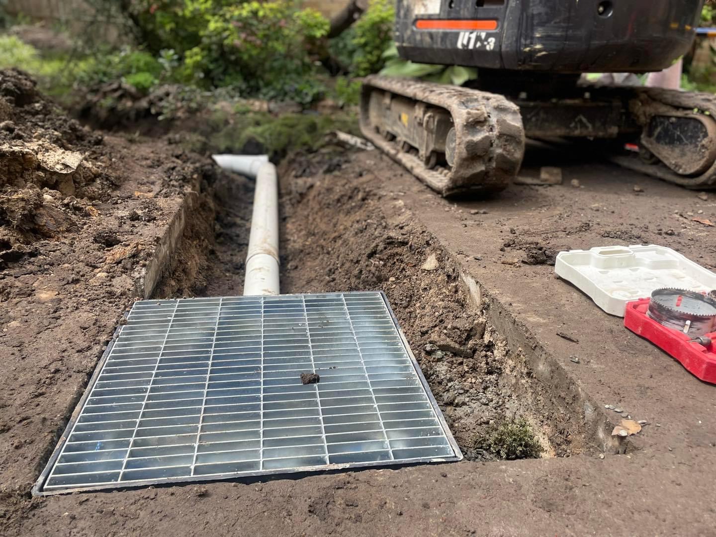 The image depicts a section of ground that has been excavated, revealing a trench with a newly installed grated drain cover on top. A large machinery, likely an excavator, is positioned nearby, suggesting that this could be part of a plumbing or drainage installation project. The grey drain pipe runs towards the grate, indicating that it is designed to collect and manage wastewater or stormwater efficiently. The surrounding earth is disturbed, indicating recent construction work, which is typical for plumbing projects such as installing or upgrading drainage systems. This installation would enhance water flow and prevent flooding in the area, showcasing the vital role of Penrith City Plumbing Services in maintaining effective plumbing systems within their service areas.
