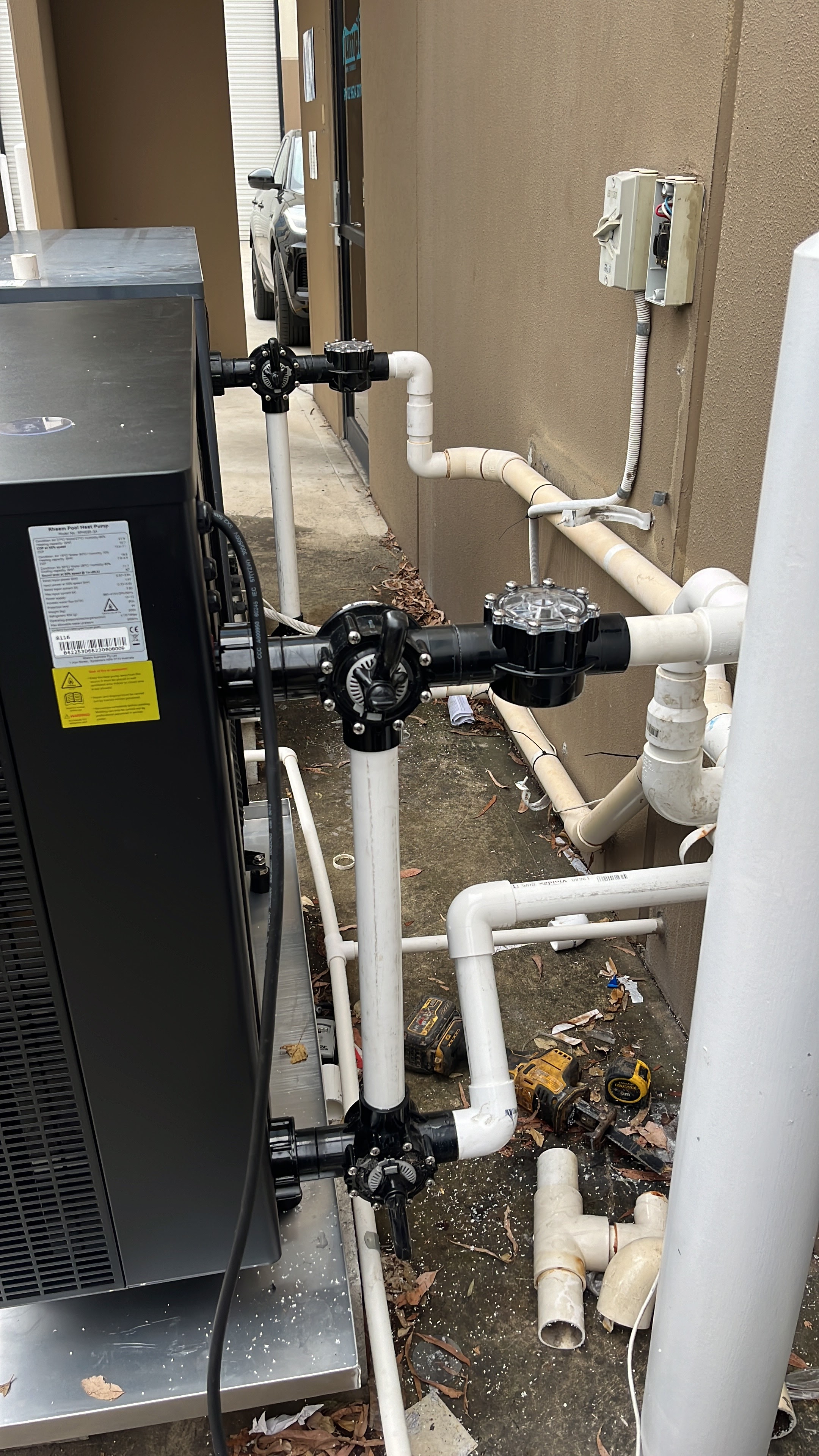 The image showcases a plumbing setup outside a building, likely in the vicinity of Penrith City Plumbing Services' operational area. In the foreground, a large black unit connected to white PVC piping is visible. The pipes are fitted with black valves, suggesting a system designed for either water supply or drainage. Nearby, there is an electrical panel, indicating the integration of electrical and plumbing systems, which is essential for operational efficiency. The area appears to be a maintenance or installation site, highlighting the technical expertise required by plumbers when dealing with complex systems. This setup reflects the types of service Penrith City Plumbing Services provides, including installation, maintenance, and repair of plumbing systems in the Mulgoa and broader Penrith areas.