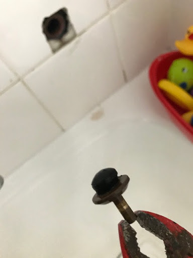 The image depicts a close-up view of a plumbing component, specifically a small brass fitting with a black rubber seal or cap on top, being gripped by a pair of pliers. The background shows a tiled wall with a visible hole, indicating a possible plumbing repair or installation site. The presence of water-related toys in a red basket suggests that the setting is likely a bathroom. This scene may be related to TBH Plumbing Solution Pty Ltd addressing a plumbing issue such as a leak or a fixture repair in the Cambridge Park area. The use of tools and the visible repairs indicate an active plumbing job, crucial for maintaining proper water flow and addressing any potential issues within the household plumbing system.