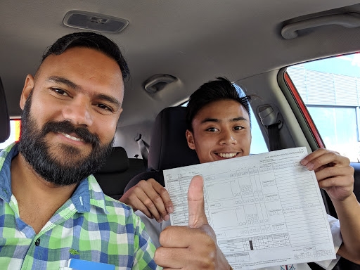 The image features two individuals seated inside a car. One person has a beard and is wearing a green and white checkered shirt, while the other is holding a document and giving a thumbs up. They appear to be celebrating a successful moment, perhaps related to driving lessons or a driving test.