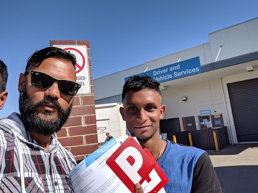 The image features two individuals standing outside a building with a sign that reads 'Driver and Vehicle Services'. One person has a beard and is holding a folder with a red 'P' sign, indicating a provisional driver's licence. The other person appears to be smiling, also holding documents. The background shows a clear blue sky and a commercial building.