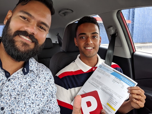 A cheerful selfie featuring two young men inside a car. One man, smiling broadly, has a thick beard and wears a patterned shirt. The other man, also smiling, holds a driving test result and gestures with a thumbs-up. The interior of the car is visible, along with the passenger seat and part of the dashboard. It appears to be a celebratory moment following a driving test.