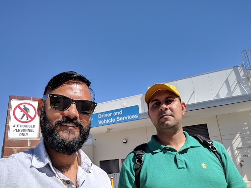 The image shows two men standing in front of a building with a clear blue sky above. One man has a beard and sunglasses, while the other is wearing a green polo shirt and a yellow cap. The building has a sign indicating it is related to 'Driver and Vehicle Services.'
