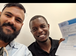 The image features two individuals, both smiling as they take a selfie together. One person holds a document that appears to display a printed score or completion sheet, indicating a successful milestone or achievement. The setting is casual and friendly, suggesting a celebratory moment.