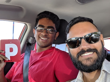 The image features two young men seated in a car. One individual is wearing glasses and a red t-shirt while holding a red letter 'P', indicating they are a provisional licence holder. The other person, with a beard and sunglasses, is smiling, suggesting a friendly and supportive atmosphere. The interior of the car shows typical driving controls, and the setting appears to be casual and relaxed.