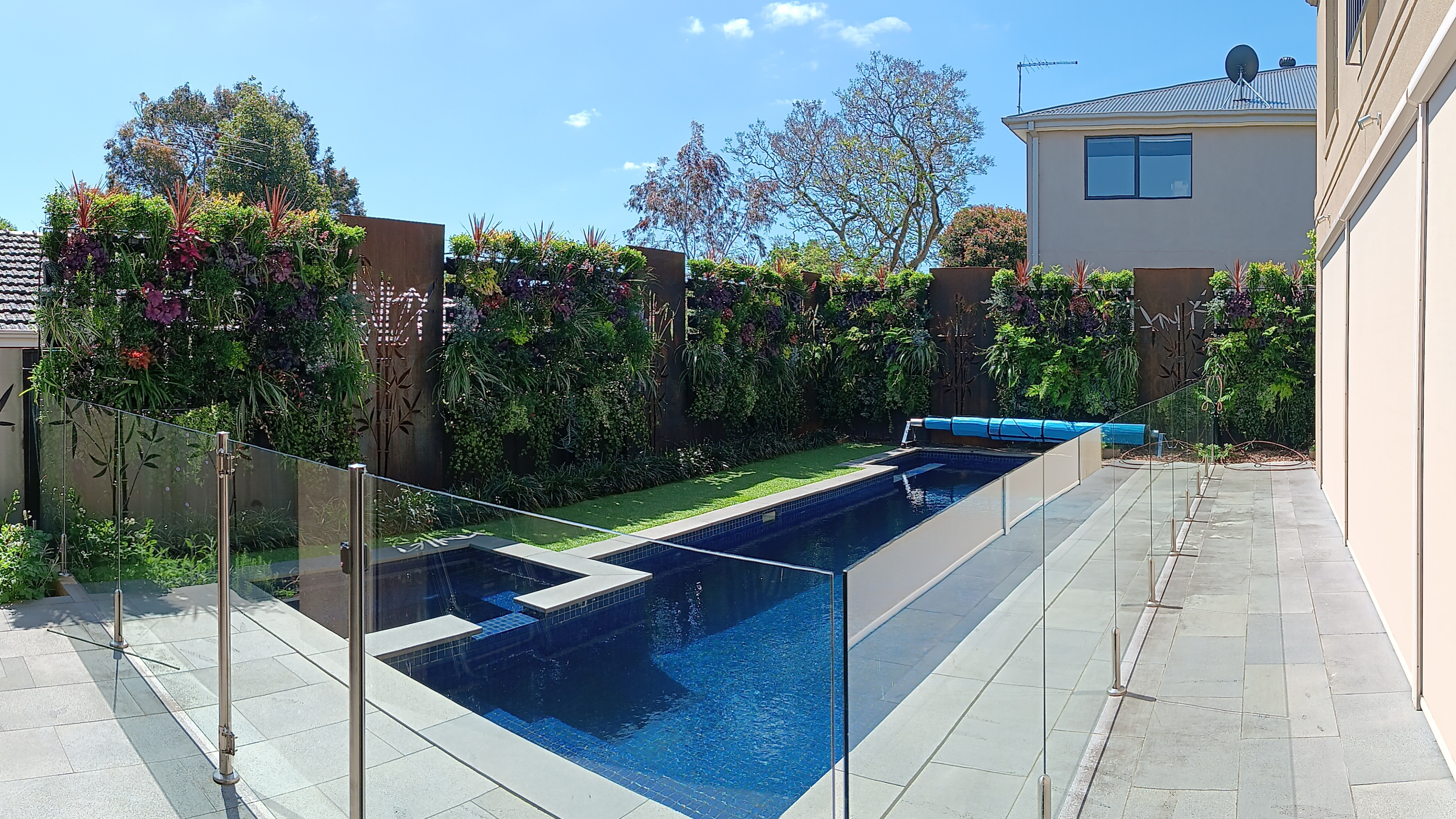 The image showcases a modern landscape design featuring a stunning swimming pool surrounded by lush green vertical gardens. The sleek, rectangular pool is bordered by minimalistic glass fencing for safety without obstructing the view. The vertical gardens consist of various plants that create a vibrant and inviting atmosphere, enhancing the outdoor space. A paved area next to the pool provides a clean, contemporary finish, while the blue water of the pool reflects the clear sky above, indicating a well-lit and pleasant environment, ideal for relaxation and leisure. This design highlights the expertise of Vertical Garden World in creating harmonious and functional outdoor living spaces.