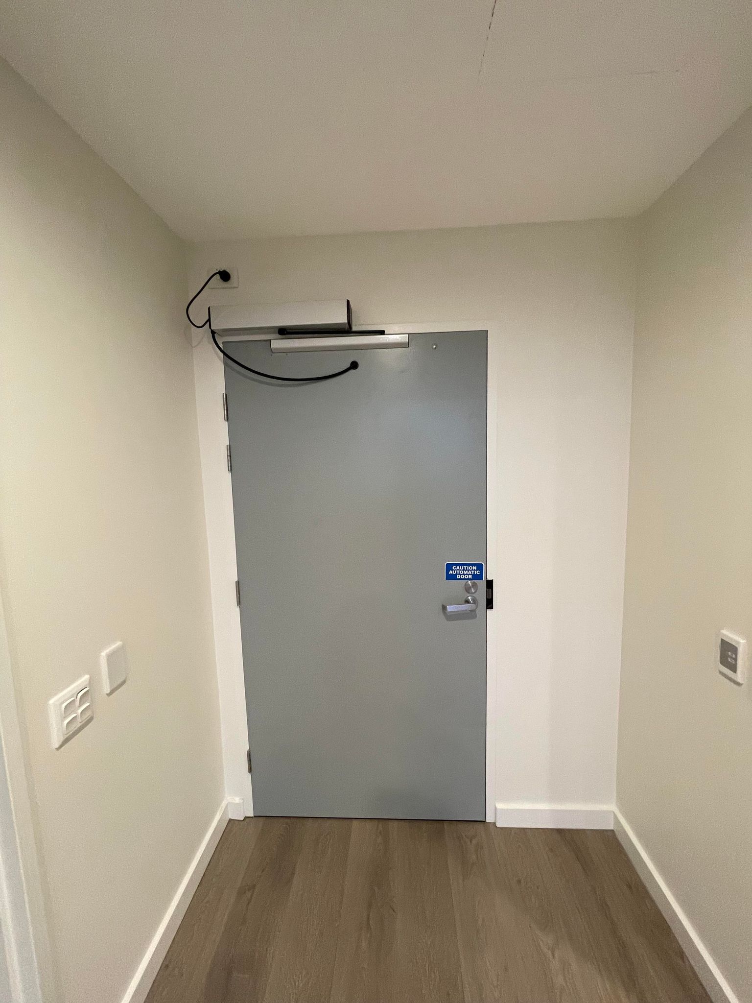 The image shows a plain corridor featuring a grey door, which appears to be a heavy-duty entrance typically used in commercial settings. The door includes a secure locking mechanism and a sign, possibly indicating emergency or access guidelines. Above the door, there's a cable connected to a device, likely part of a security system. The flooring is wooden, giving a modern touch to the space. In the context of SJC Building & Construction, this image could represent recent renovations or new constructions they are involved in, focusing on enhancing safety and security in commercial buildings, crucial for areas frequented by the Australian Defence Forces.