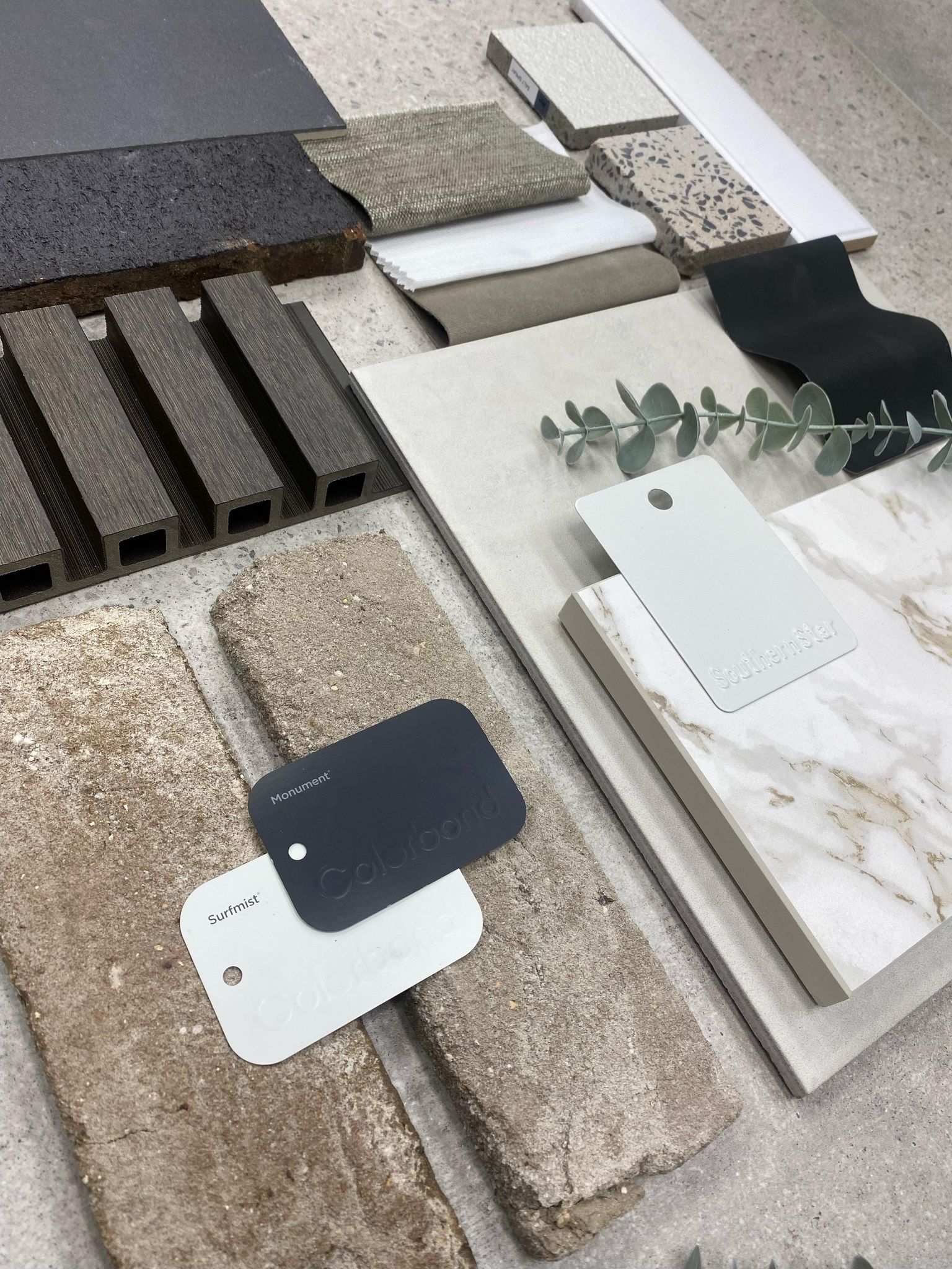 The image features a meticulously arranged collection of building materials and design samples on a textured surface. Prominent are various tiles and slabs, showcasing a mix of textures including smooth, matte, and polished finishes. The colour palette includes earthy tones, soft whites, and modern blacks, reflecting contemporary architectural trends. Sample cards, labelled with different textures and colours, are positioned prominently, suggesting a design consultation or selection process for a construction project. This visual presentation indicates that SJC Building & Construction is engaged in the early stages of a project, possibly gathering materials for residential or commercial construction in their serviced areas.