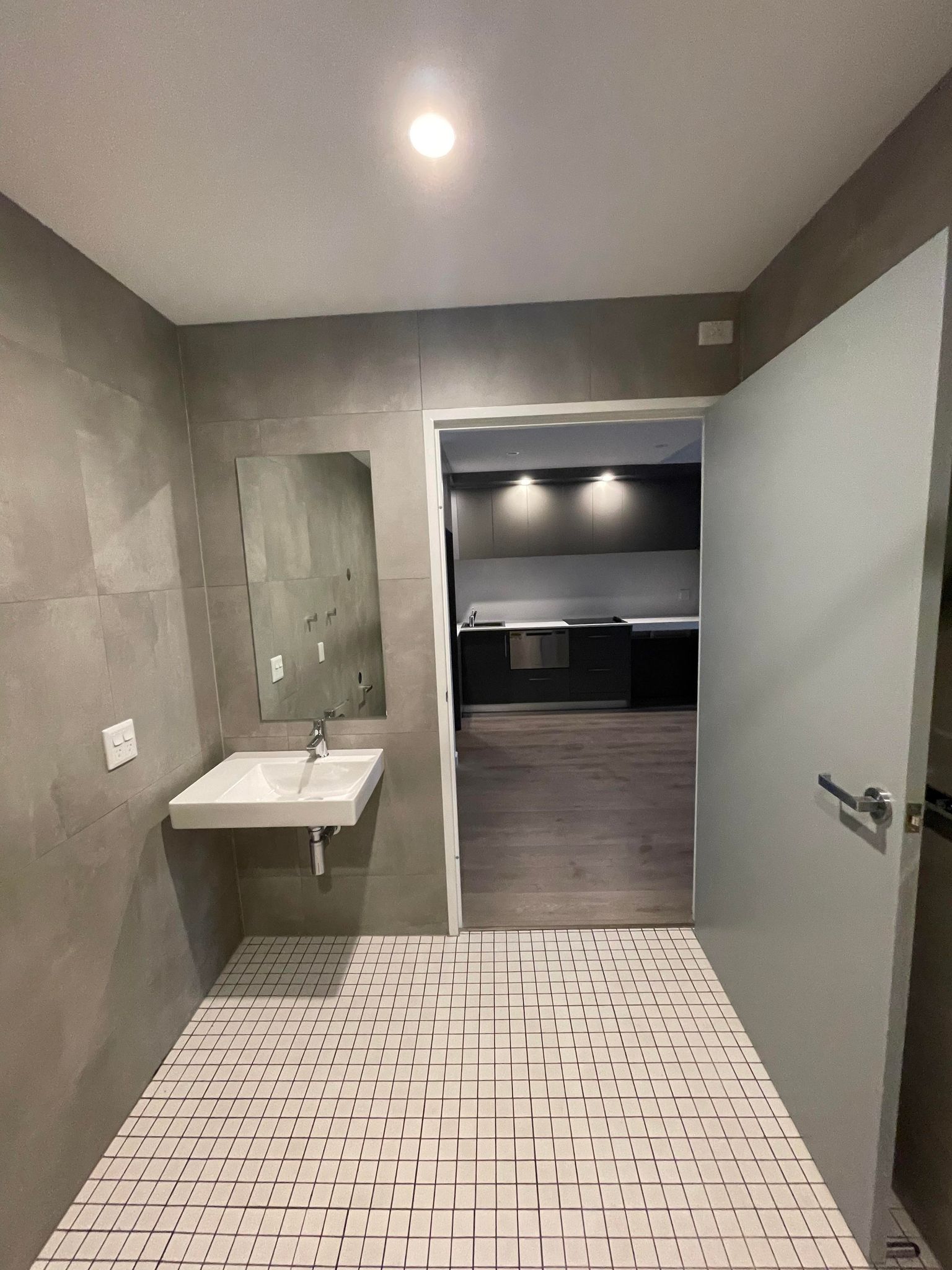 The image shows a modern bathroom interior featuring a minimalist design with grey tiled walls and a grid-patterned floor. There is a sleek, wall-mounted sink with a single tap, and a mirror positioned above it, reflecting the clean lines and contemporary aesthetic typical of SJC Building & Construction projects. The room is well-lit with recessed lighting, highlighting the functional space. Beyond the bathroom, an open doorway reveals a kitchen area that appears spacious and modern, but is partially out of frame. This layout suggests an ongoing construction or renovation project, aligning with SJC's focus on creating stylish and functional spaces for residential and commercial clients. Attention to detail and quality is evident in the choice of materials and the overall design.