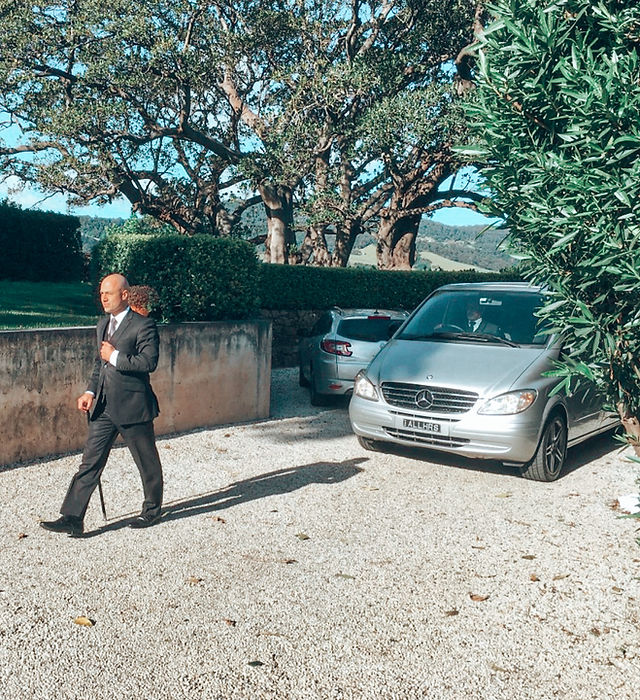 In the image, a well-dressed individual is walking along a gravel path, likely preparing for a transportation service engagement. He holds a cane, suggesting he is either a client or a representative of All Hours Transfers, demonstrating professionalism and confidence. Behind him, a silver Mercedes van is parked, indicative of a luxury transport offering. The setting appears serene and upscale, with robust greenery framing the area, which is typical for the transport services catering to Inner Sydney and surrounding suburbs. The ambiance suggests that All Hours Transfers focuses on providing comfortable and upscale transport solutions to clients in Kingsgrove and the Eastern Suburbs.