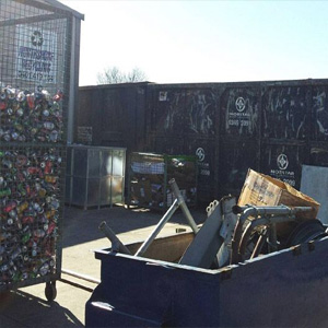 The image shows an outdoor scrap metal yard with various containers filled with scrap metal and crushed cans. One container holds compressed aluminium cans, while another contains assorted metal parts and scrap items. The facility appears secure and organised, with signage featuring the Transformer Metals logo visible on the containers and fencing in the background. The setting is sunny with clear skies, reinforcing a professional metal recycling environment.