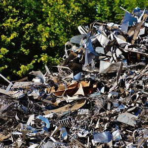 The image shows a large pile of scrap metal consisting of various metallic parts, including sheets, wires, and machinery bits, reflecting the typical collection at a metal recycling yard. The background features green foliage, indicating the pile is outdoors, likely at a scrap metal dealer site such as Transformer Metals in North Geelong, VIC.