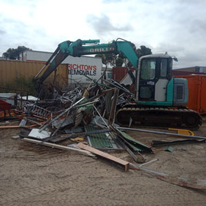 The image shows a scrap metal yard with a turquoise excavator lifting and sorting a large pile of mixed scrap metal, including rusted sheets and metal rods. The ground is dirt, and a container and signboard for a removal service are visible in the background. The scene represents active scrap metal processing, typical for a metal recycling business in North Geelong, VIC.