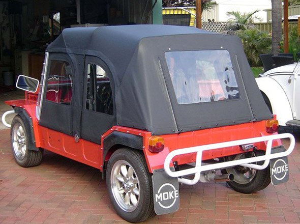 The image showcases a vintage-style vehicle known as a Moke. It's a small, convertible car with a bright red body and a black, soft-top roof. The rear view highlights its unique design, featuring a clean exterior with modern alloy wheels. The car is parked on a brick surface in what appears to be a garage or workshop setting, indicating it may be in the process of restoration or renovation. The presence of the logo "MOKE" on the rear adds a touch of nostalgia, appealing to car enthusiasts. This vehicle could be a perfect project for Horgans Auto Upholstery, as they specialise in restoring and enhancing classic cars.