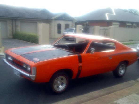 The image features a classic orange car parked on a residential street. The vehicle has a sleek design, with a distinctive curved roofline and a bold paint job, highlighting its vintage appeal. Its black racing stripes along the hood and side add an aggressive touch to the overall appearance. The backdrop shows a suburban setting with modern houses and neatly maintained driveways, indicating a well-kept neighbourhood. This car, possibly a muscle car, represents a type of vehicle that car restoration enthusiasts at Horgans Auto Upholstery may specialise in restoring, bringing classic models back to life with quality craftsmanship.