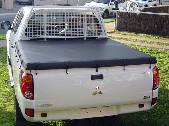 The image showcases a Mitsubishi Triton utility vehicle, featuring a black tarp cover on its tray. The cover is secured with fasteners, indicating its purpose to protect cargo. A protective mesh screen is also visible behind the cab, suggesting a focus on vehicle functionality and safety. The truck is parked in a residential area, hinting at its usage for both personal and work purposes.