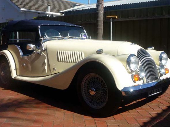 The image features a classic vehicle, elegantly styled, with a cream-coloured body and a black convertible hood. The car is parked on a brick-paved driveway, surrounded by a residential area, which enhances its vintage appeal. The vehicle showcases chrome details, including headlights and a grille, accentuating its restoration quality. The pristine condition of the car suggests meticulous care and maintenance, typical of a vehicle restored by Horgans Auto Upholstery. This setting reflects the character of an auto restoration service that values both aesthetics and craftsmanship.