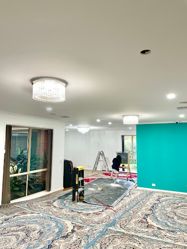 The image depicts an interior space, likely a residential setting, with freshly installed lighting fixtures creating an inviting atmosphere. Two chandeliers are visible, adding elegance to the ceiling. A ladder indicates that work is currently being completed, possibly involving the installation of electrical fixtures or painting, as suggested by the colour on the wall. The flooring is covered with a decorative rug, while tools and materials are scattered about, signifying an active renovation or improvement process. The bright walls, especially the accent turquoise colour, contrast with the neutral tones of the remaining space, enhancing its modern aesthetic.