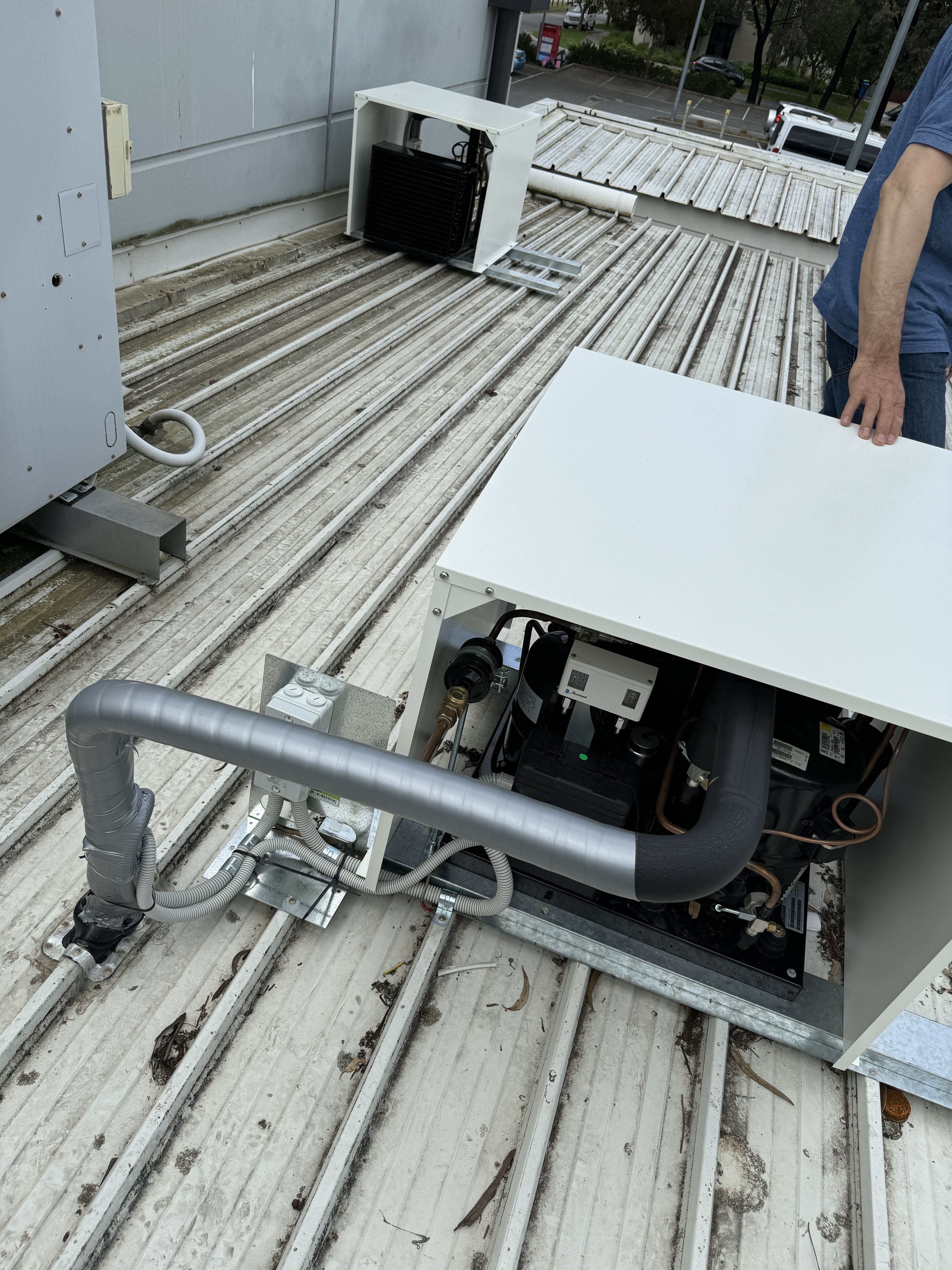 The image depicts a rooftop setup where an electrician is working on an air conditioning unit. The unit is partially exposed, revealing its internal components, including tubing and electrical connections. The surface appears to be a metal or corrugated roofing material, which suggests that this work is part of routine maintenance or installation. The presence of such equipment indicates the importance of utilising qualified electricians for technical tasks to ensure safety and compliance with regulations.