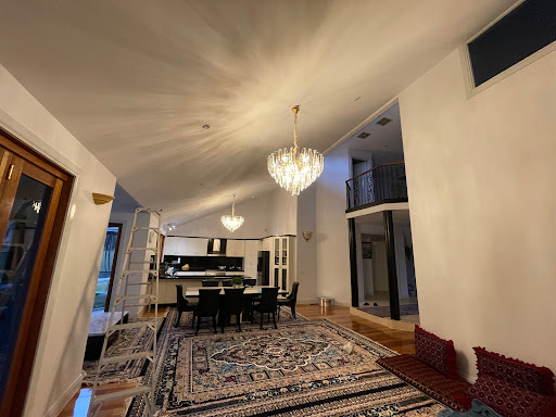 The image depicts a spacious and modern interior of a home featuring high ceilings and elegant lighting. Two crystal chandeliers illuminate the room, casting a warm glow. The flooring is polished wood, and there is a large, ornate rug providing a central focus for the living area. A dining table is visible in the background, accompanied by black chairs, suggesting a comfortable and stylish space for gatherings. To the left, a ladder indicates that recent work may have been carried out, possibly related to the lighting installation.