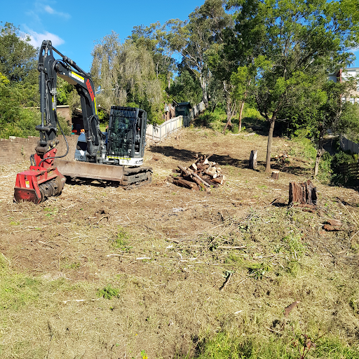 The image depicts a land clearing site, where an excavator is seen on a mounded area with a clear blue sky above. The excavator is positioned next to a pile of timber and debris, showcasing the aftermath of recent tree removal and land clearing operations. Surrounding the site are several trees, indicating a transformation of the area for better land management. The ground is uneven with patches of grass and exposed soil, typical of land preparation for further developments or maintenance. This visual reflects the services offered by Reeper Mulching in tree and land management.