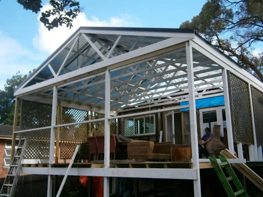 The image shows a partially constructed outdoor living space, possibly a deck or terrace, featuring a large open area with a pitched roof. The structure is framed with wooden beams and a lattice design along the lower sections, which provides both support and aesthetic appeal. In the background, there are chairs and tables, suggesting the space is designed for relaxation and social gatherings. A worker can be seen, contributing to the build. The setting looks serene, surrounded by trees and a clear sky, indicating a desirable outdoor environment.