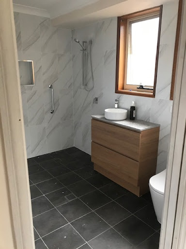 The image depicts a modern bathroom featuring a sleek design with a shower area, a wooden vanity with a stone countertop, and a wall-mounted basin. The tiles are laid in a contemporary pattern, offering a clean and stylish look. Natural light is filtered through a window, enhancing the overall ambiance. The bathroom is fitted with necessary fixtures including a towel rail and a toilet, showcasing an elegant balance of functionality and aesthetics.