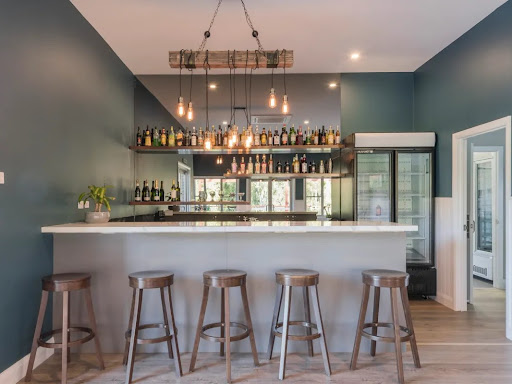 The image features a modern, stylish home bar with a spacious countertop and high stools that complement the design. The walls are painted in a deep green, enhancing a contemporary and inviting atmosphere. Above the bar, a unique lighting fixture made from wood adds an industrial touch while providing warm illumination. Behind the bar, an assortment of liquor bottles is neatly arranged on shelves, indicating it's well-stocked for entertaining. The overall design reflects a blend of sophistication and comfort, ideal for residential spaces.