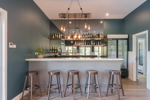 The image showcases a modern home bar area with a clean and contemporary design. The bar features a sleek countertop with a minimalist aesthetic, complemented by several high stools, offering an inviting space for social gatherings. Behind the bar, an impressive collection of bottles is displayed prominently on shelves, highlighted by warm pendant lighting, adding to the overall ambience. The walls are painted in a rich dark blue, creating a stylish contrast with the light wood flooring. This space reflects a perfect blend of functionality and stylish design, ideal for a residential build project.