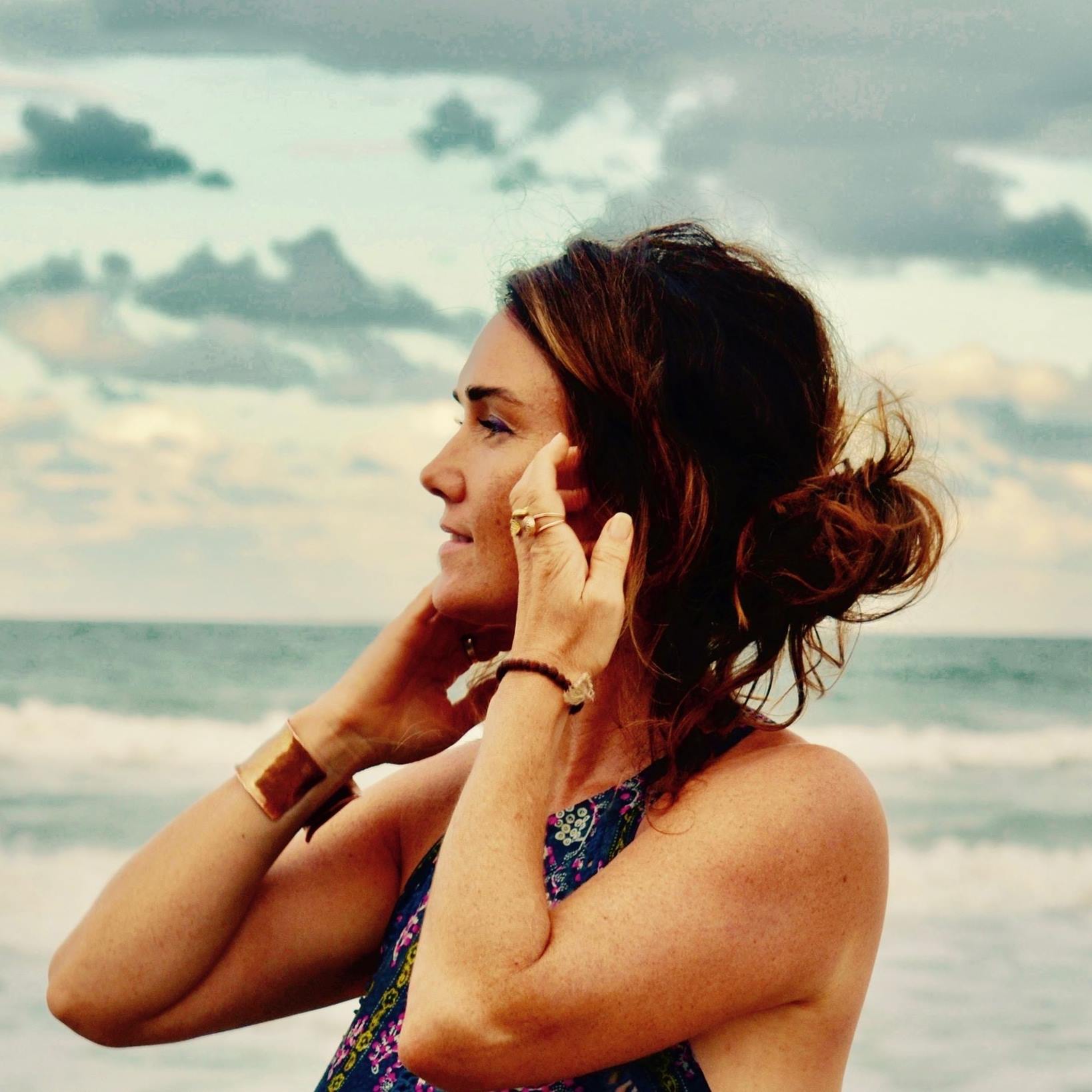 The image features a woman standing by the beach, her silhouette framed by a backdrop of a cloudy sky and the ocean. She has a relaxed posture, delicately touching her face with one hand, while her other hand is tucked near her hair, which is styled in loose waves. The scene evokes a sense of tranquillity and a connection to nature, reflecting the ethos of naturopathy. The setting could be used to promote the natural, holistic approach of the Herb Bar, suggesting harmony with the environment and self-care practices.