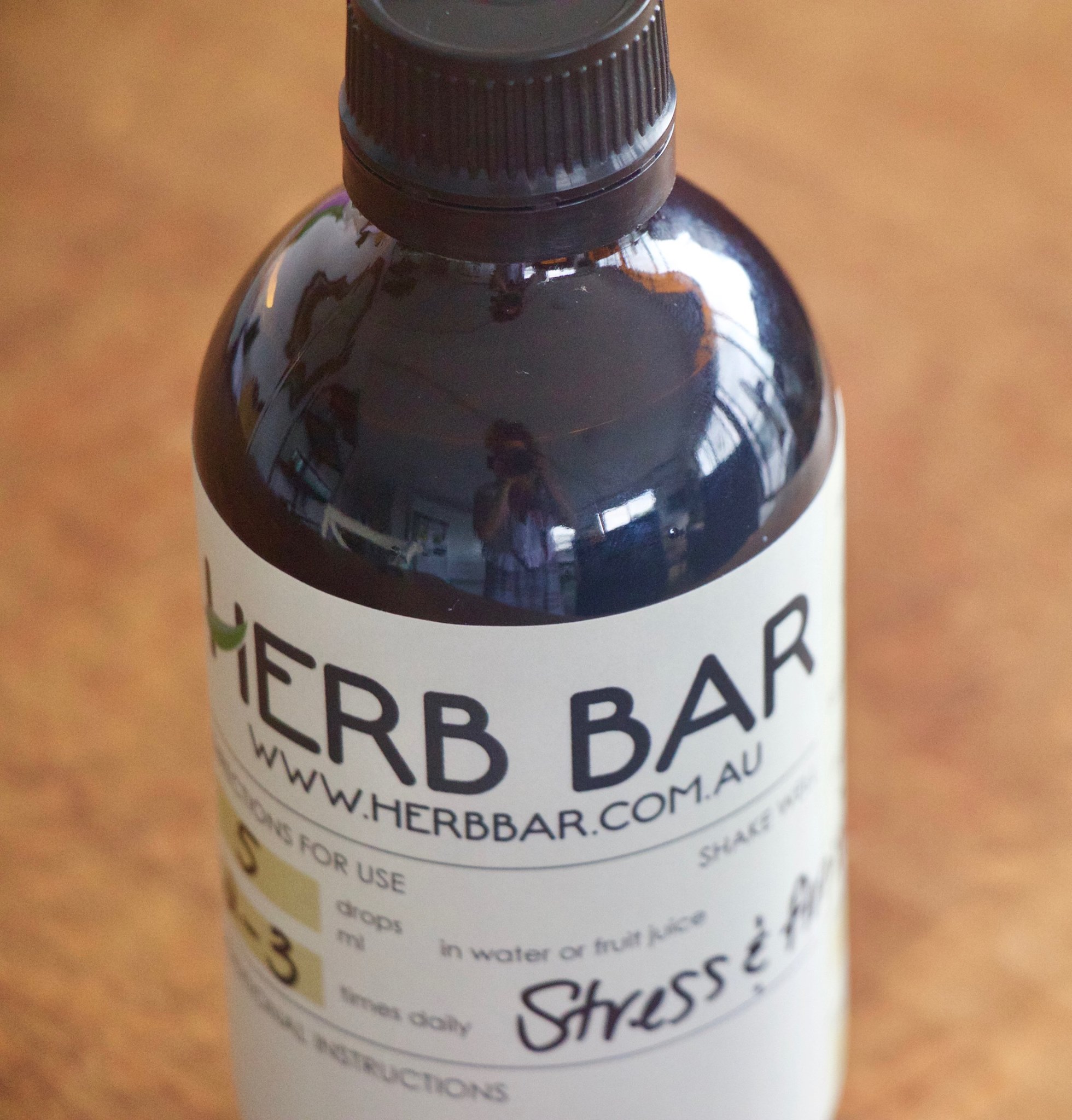 The image showcases a bottle labelled as part of the Herb Bar, a Naturopath business. The bottle has a dark, opaque design, which helps maintain the integrity of its contents, usually believed to be herbal extracts or remedies. The label features the business name 'HERB BAR' prominently, along with the website URL 'www.herbbar.com.au'. The instructions indicate to shake the bottle and specify dosage as '5 drops' to be mixed with water or fruit juice, to be taken three times daily. The highlighted purpose of this particular extract is to address 'Stress & Anxiety', indicating its therapeutic use. The background suggests a warm, natural setting, likely emphasising the naturopathic and holistic focus of the business.