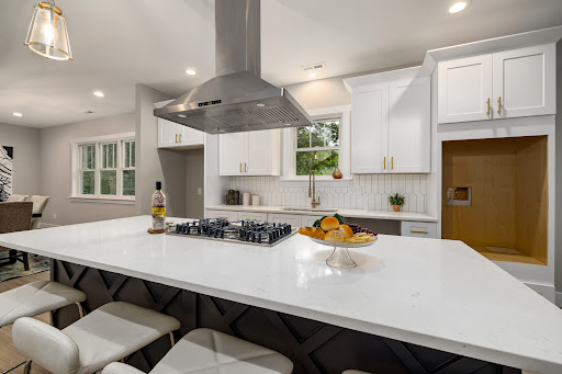The image showcases a modern kitchen renovation, indicative of high-quality work that Accent Ducting offers. The kitchen features a sleek, spacious design with a large central island, topped with a pristine white countertop that pairs beautifully with the light cabinetry. A contemporary hood vent hangs above a professional-grade cooktop, emphasising functionality and style. On the island, a decorative bowl filled with fresh fruit adds a vibrant touch, promoting a welcoming atmosphere. The cabinetry is neatly arranged, providing ample storage and an organised aesthetic. Soft, ambient lighting illuminates the area, creating a warm and inviting feel. This modern design reflects the expertise and attention to detail that Accent Ducting is known for in their kitchen renovation projects, appealing to homeowners in Brisbane and surrounding areas.