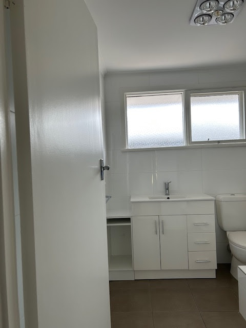 The image depicts a well-lit, modern bathroom featuring a clean design. The bathroom includes a white vanity with multiple drawers and a sleek sink, alongside a white toilet. A large window allows natural light to fill the space, highlighting the tiled walls and floor. The overall aesthetic is simple and functional, suggesting a recent renovation or maintenance work, ideal for showcasing the quality service provided by Bellarine Painting and Property Maintenance.
