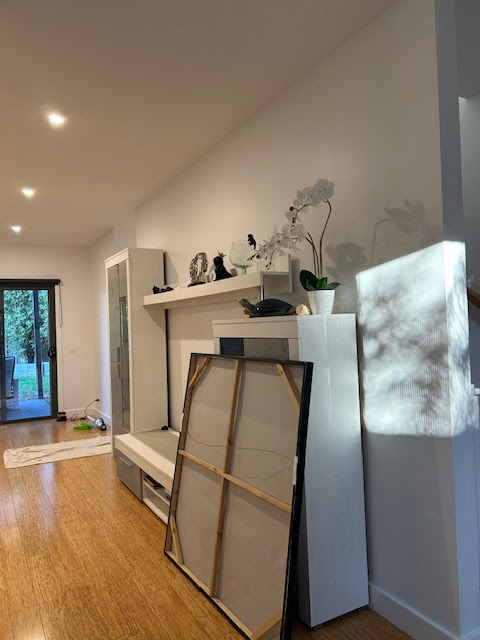 The image depicts a modern interior space, possibly a living room or foyer, characterised by clean lines and a minimalist aesthetic. To the left, a large white shelf unit stands against a wall, adorned with decorative items and a small plant. The floor is made of bamboo, indicating a warm and natural finish. In the foreground, a framed picture leans against the shelf, suggesting ongoing organisation or decorating efforts. Soft lighting from ceiling fixtures provides a bright ambiance, while a window in the background invites natural light and offers a glimpse of an outdoor space, possibly a garden or patio. This contemporary area reflects the principles of space management and aesthetic appeal, relevant to property maintenance and painting services.