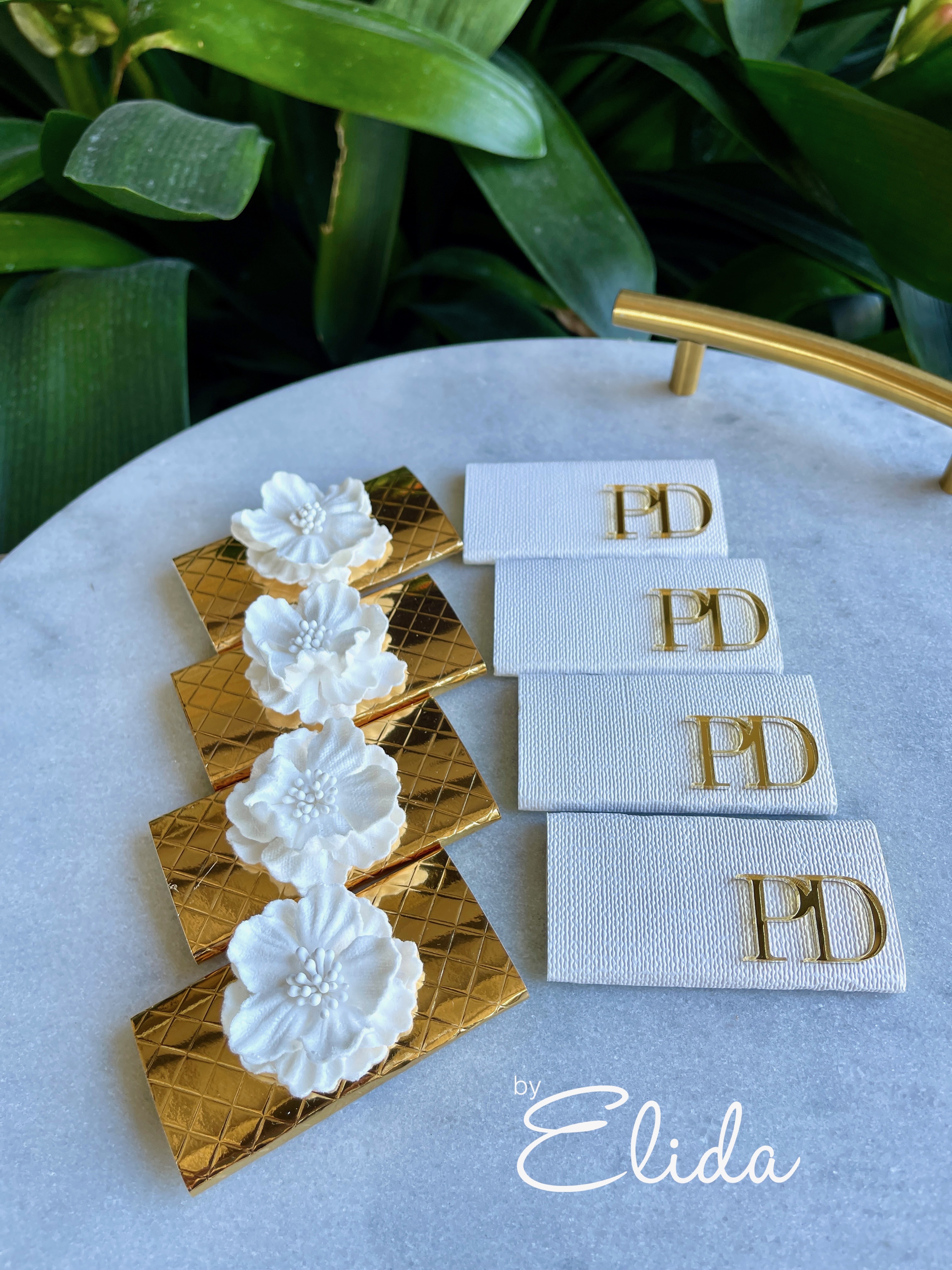 The image showcases elegant wedding stationery or place cards designed by By Elida. There are four distinct designs: two in a luxurious gold finish featuring textured patterns and delicate white floral embellishments, adding a touch of sophistication. The other two cards are in a pristine white, each adorned with a subtle gold monogram ‘ED’, representing the brand. The backdrop of lush green leaves complements the refined aesthetic of the stationery, setting a serene and stylish tone suitable for weddings.