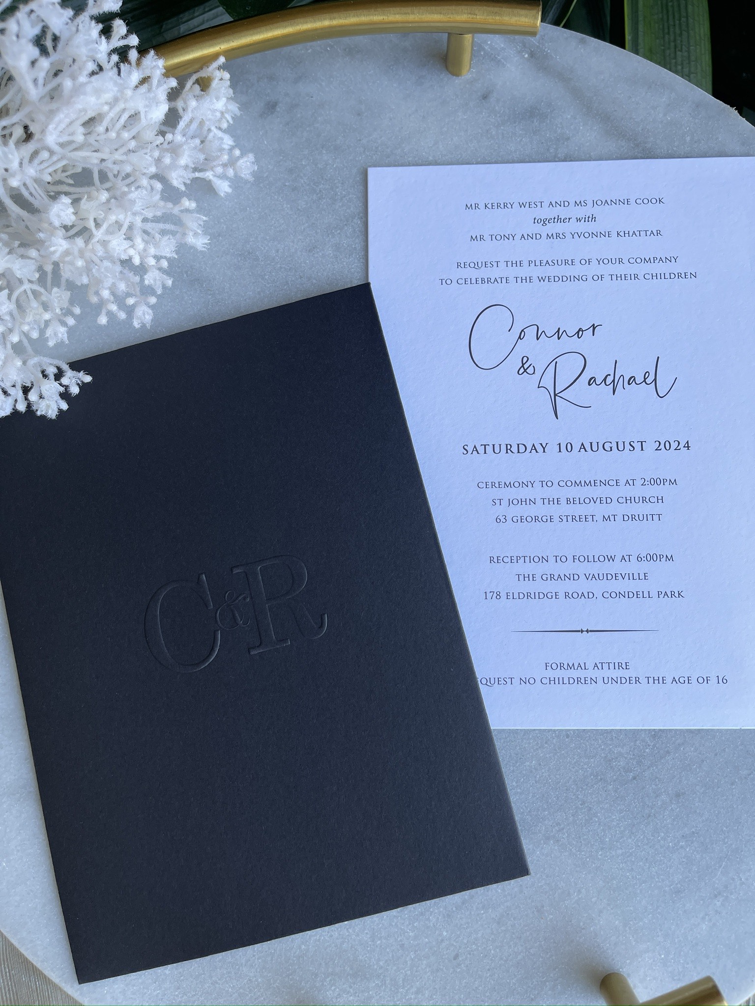 The image features a sophisticated wedding invitation suite for Connor and Rachael's wedding, dated for Saturday, 10 August 2024. The invitation is elegantly printed on quality paper, highlighting a stylish embossed logo featuring the initials 'C' and 'R'. It includes details about the ceremony at St John the Evangelist Church and the subsequent reception at The Grand Vaudeville. The overall aesthetic is refined and formal, suggesting a classy wedding theme, with a note indicating proper attire and a reminder for guests regarding children attending.