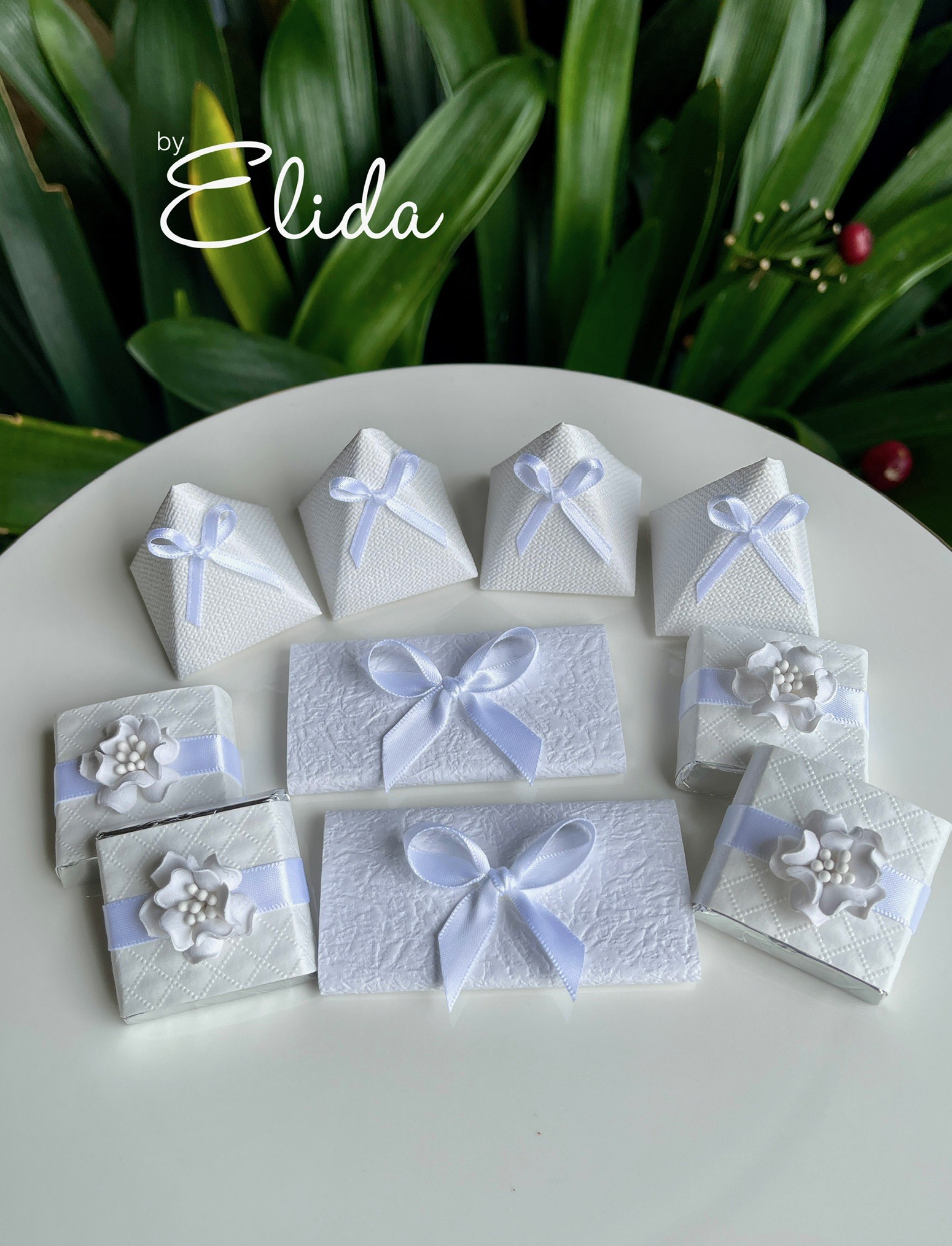 The image showcases beautifully crafted wedding favours designed by Elida. Arranged elegantly on a white plate, there are an assortment of small boxes and packages. These include pyramid-shaped boxes and flat rectangular boxes, all in a pristine white finish, tied with delicate pastel blue ribbons. Some packages feature intricate floral decorations, giving them a romantic touch. The backdrop of lush green leaves adds a natural and inviting feel, perfect for a wedding setting. This presentation highlights the elegance and attention to detail that Elida brings to wedding services in Canada Bay.