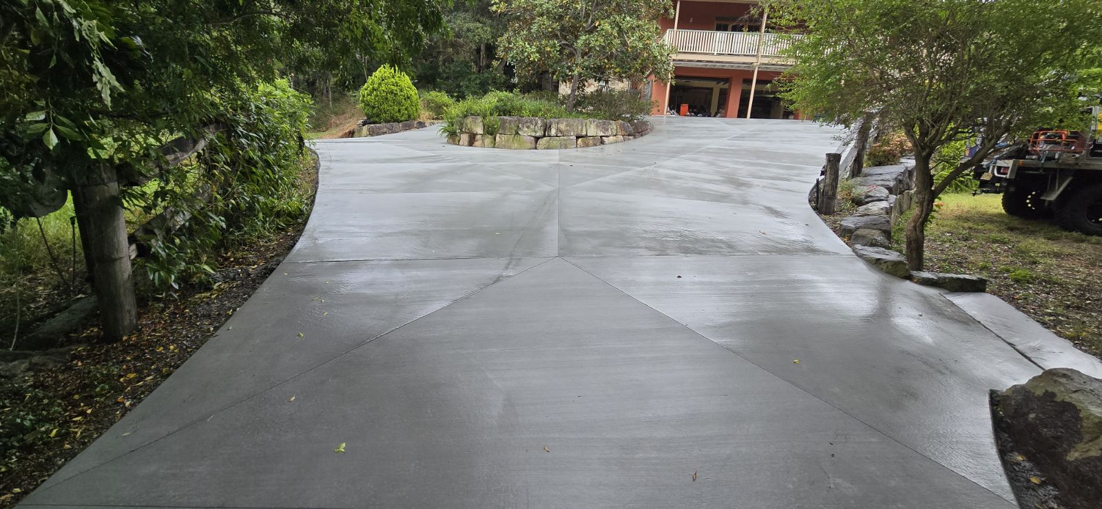The image showcases a freshly completed concrete driveway, featuring a smooth and polished surface. The driveway is designed with a gentle curve and intersecting lines that create an appealing visual pattern, indicative of professional concrete work. Lush green foliage surrounds the driveway, enhancing the ambience and showcasing how well the concrete integrates with the natural landscape. The driveway appears to lead up to a residential property, suggesting a well-designed entrance that adds value to the home.