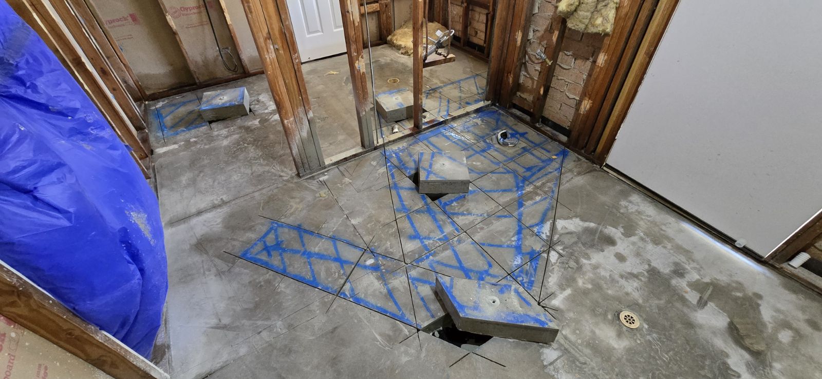 The image shows a concrete floor that has been partially cut and marked for further work. Blue tape outlines specific areas for cutting or repair, indicating precise measurements and planning for plumbing or drainage installations. The surrounding walls are exposed, suggesting that this is part of a renovation or construction project. Tools and equipment typical for concrete work might be present or implied, given the context of the business as a concrete contractor. Overall, the scene reflects a professional environment focused on precise concrete cutting and installation services.