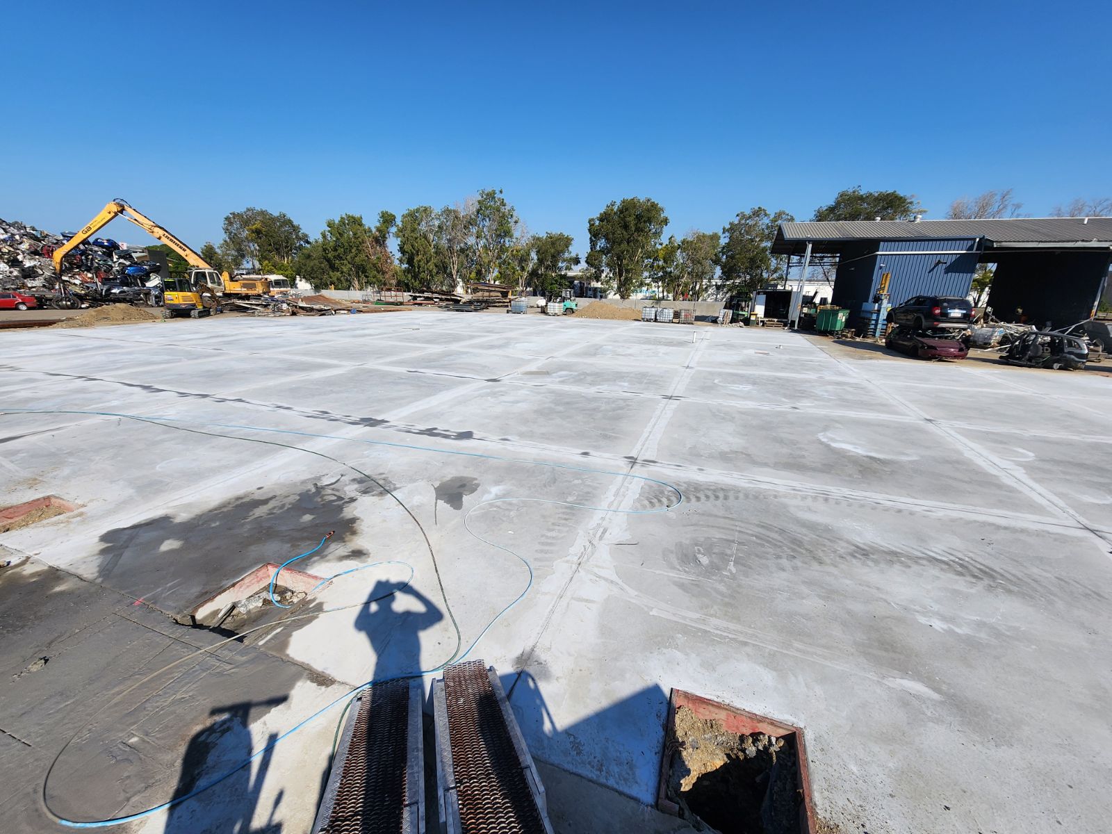 The image depicts a large, recently poured concrete slab in a construction or industrial area. The surface appears smooth and level, with some markings suggesting recent finishing work. In the background, several vehicles and machinery, including an excavator, indicate ongoing operations at the site. The area around the slab features trees and what appears to be a scrap or recycling yard, suggesting that this might be related to construction, demolition, or concrete cutting activities.